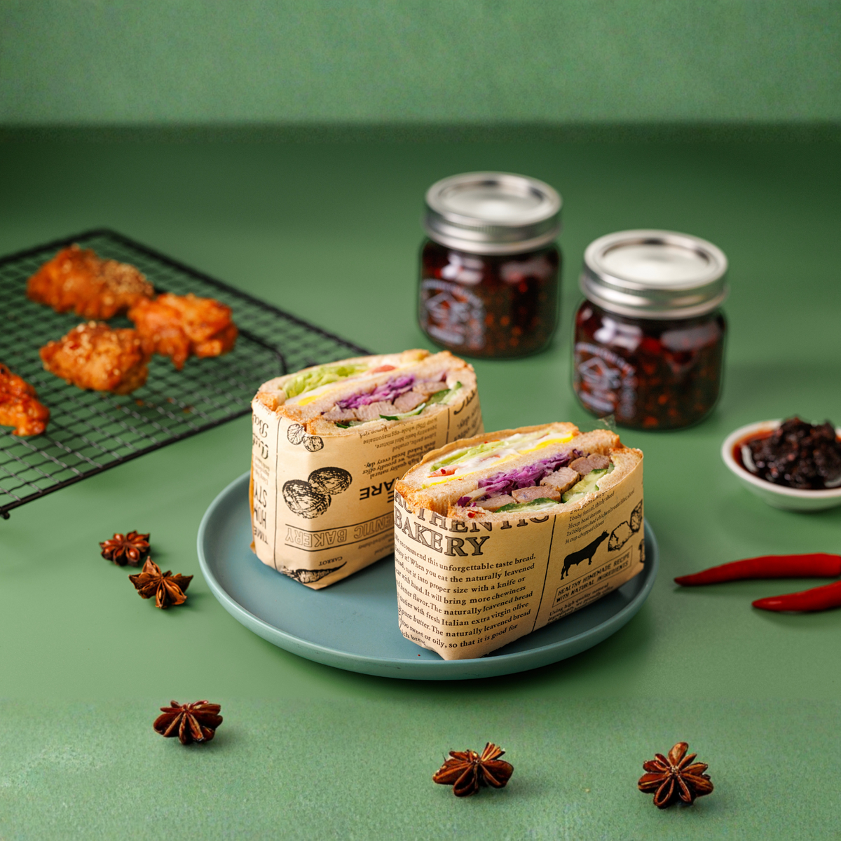 Two wrapped sandwiches on a green surface with jars and baked items in the background.