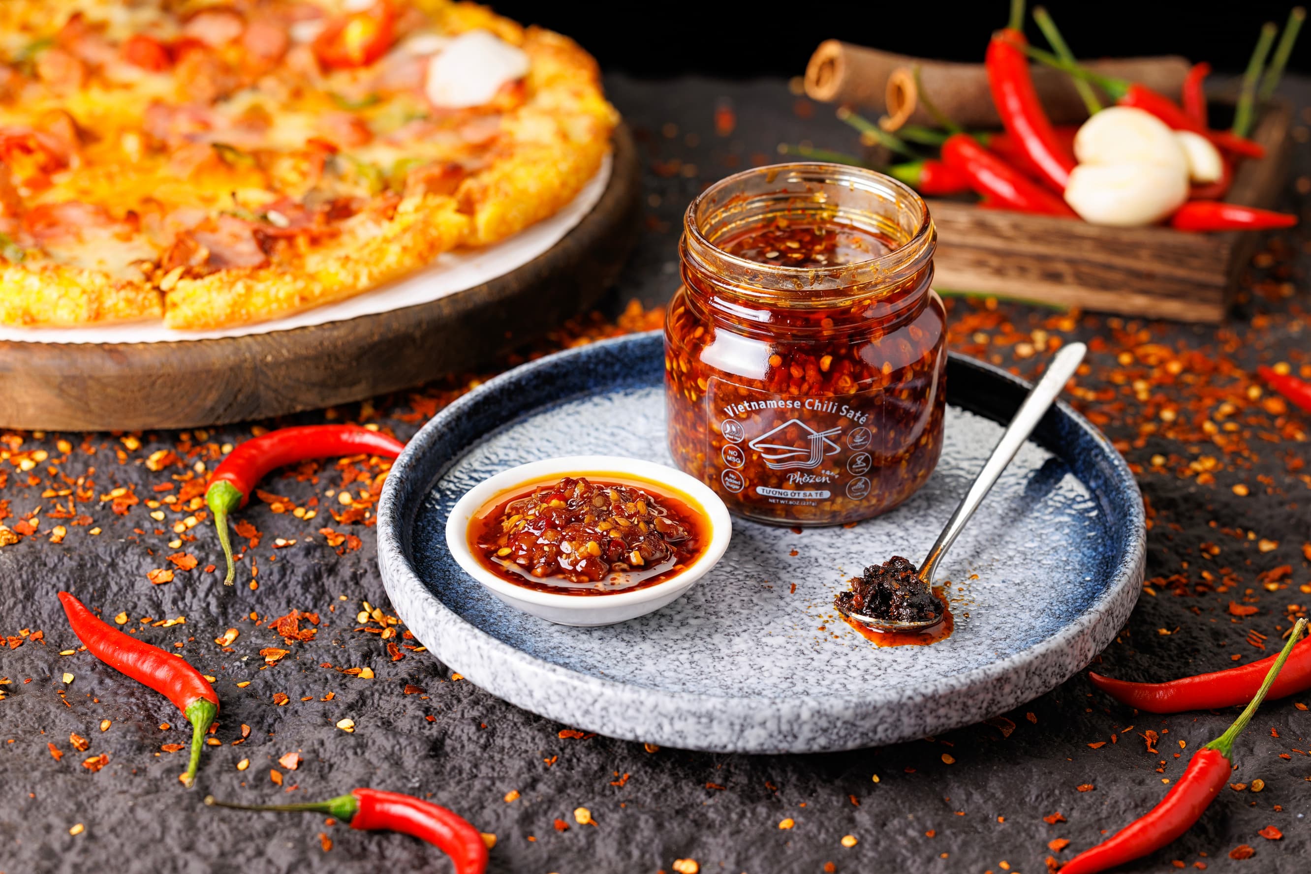 Jar of chili sauce on a plate with a pizza and red peppers in the background