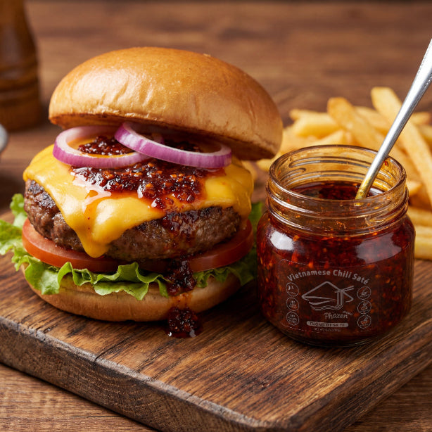 Hamburger with cheese, lettuce, tomato, and red onion on a wooden board with a jar of Vietnamese chili sauce.