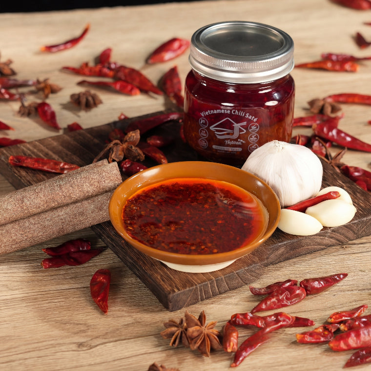 Jar of chili paste with a bowl of red sauce, surrounded by dried red chilies and star anise on a wooden surface.