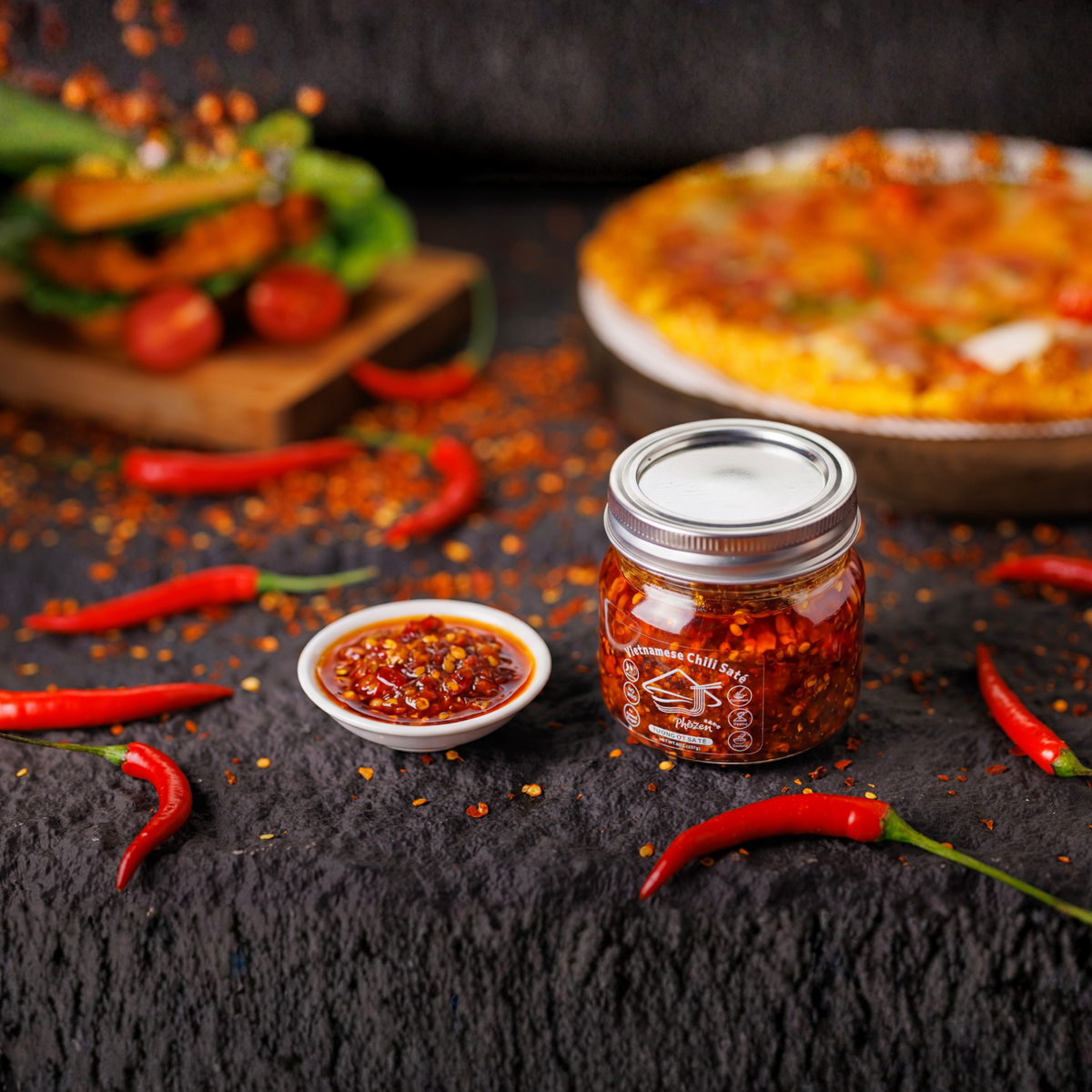 Jar of chili paste with a small bowl of chili sauce and red chilies on a dark surface.