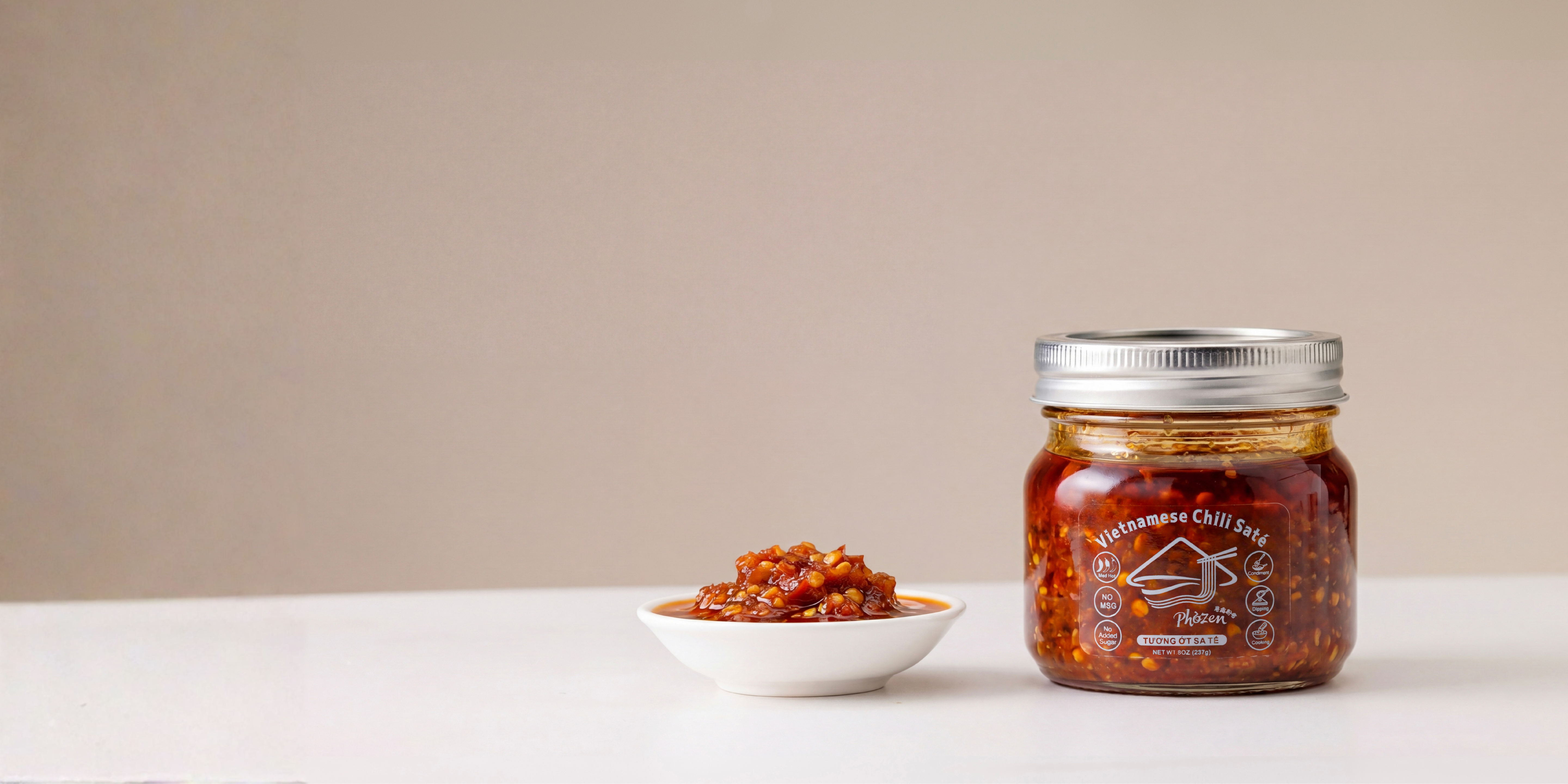 Jar of chili paste with a small bowl of chili on a white surface against a beige background