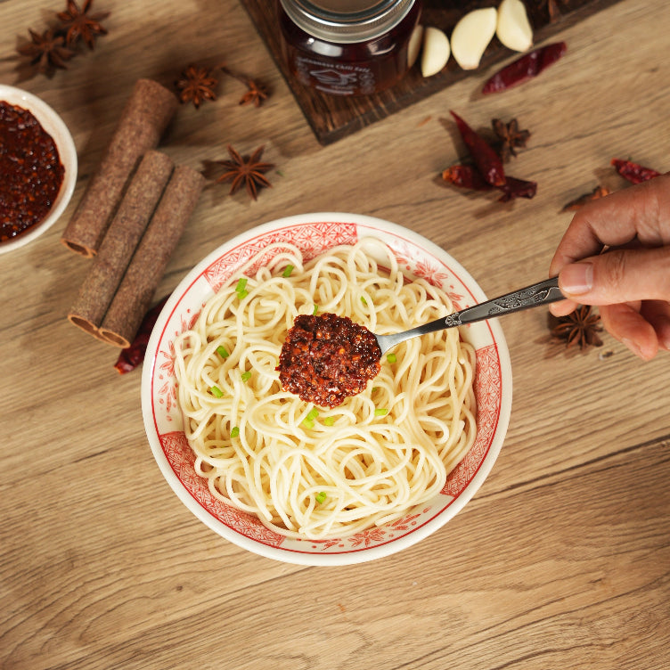 Bowl of noodles with a spoonful of red sauce on a wooden table with spices and a bottle.